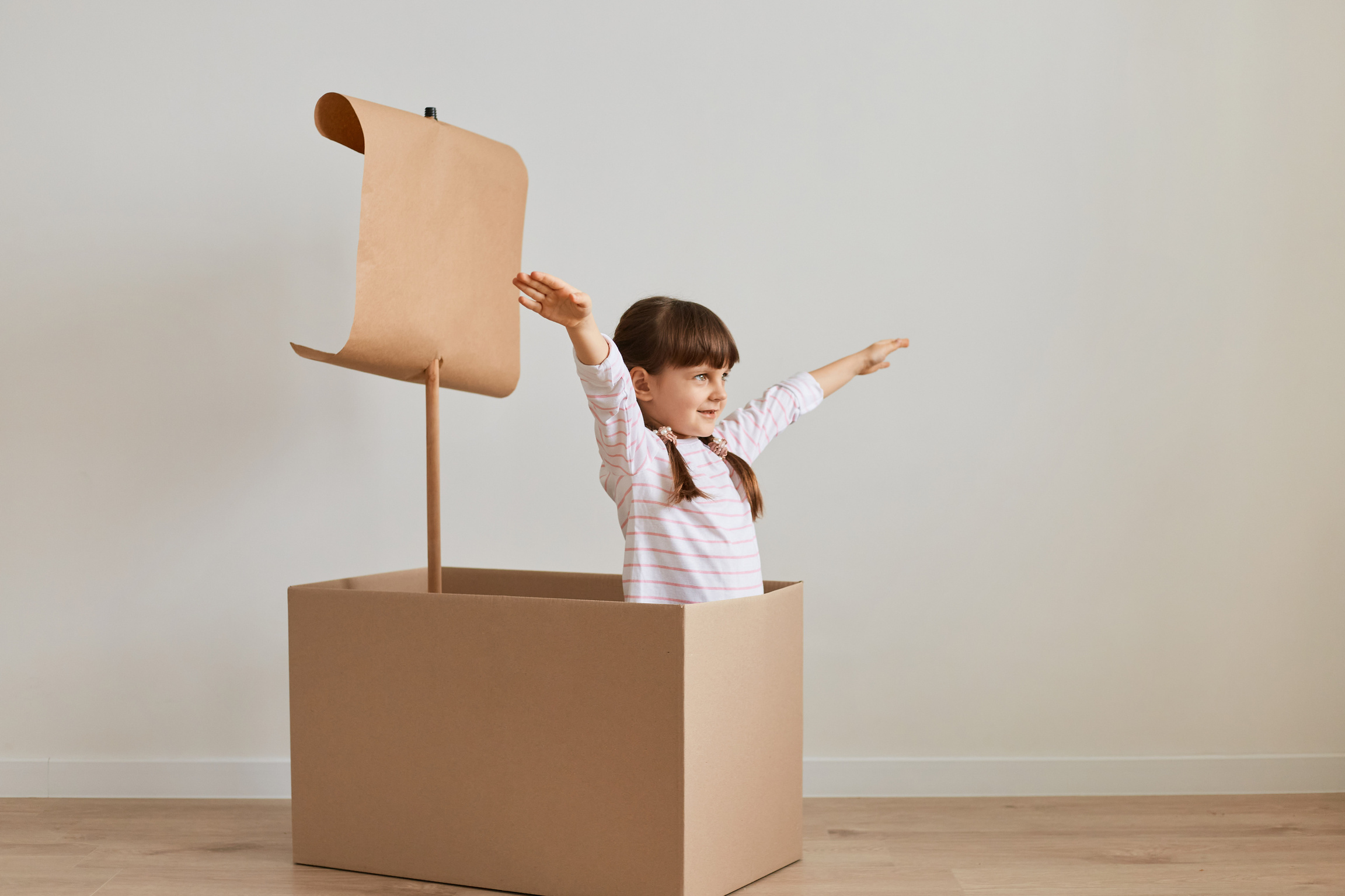 Indoor Shot of Excited Little Girl Sitting in Cardboard Box with Paper Flag, Playing Ship Capitan, Pretending She Is in the Sea, Spreading Her Hands Aside like Flying, Enjoying Her Game.